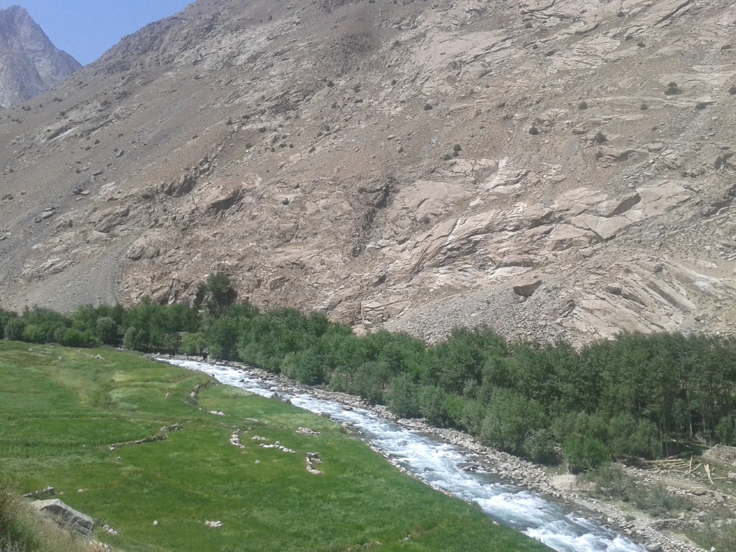 The Weghnan River flows between a green bank and trees below a rocky mountainside in Warsaj District, Takhar.